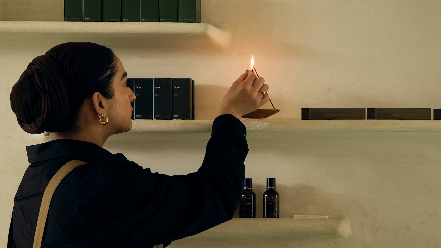 Figure lights an incense stick on a white shelf with Aesop products surrounding.