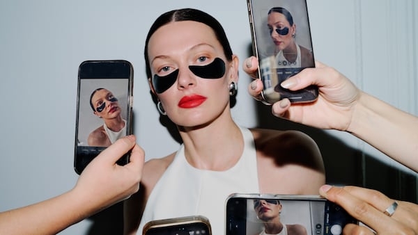 A woman with slicked-back hair and red lipstick wears black under-eye masks while several people hold smartphones taking photos of her from different angles.