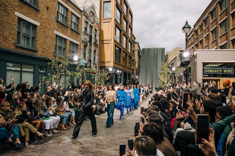 Outdoor fashion show on a narrow cobblestone street, with models walking in a line wearing David Koma designs. The front model is dressed in black leather, followed by others in blue and neutral-toned outfits. The audience sits on both sides of the runway, many holding up phones to record the event.